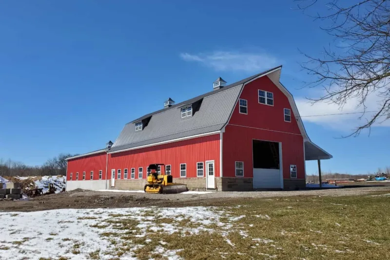 Red gambrel-roof agricultural pole barn with stone wainscot, cupolas, and a wide sliding end door