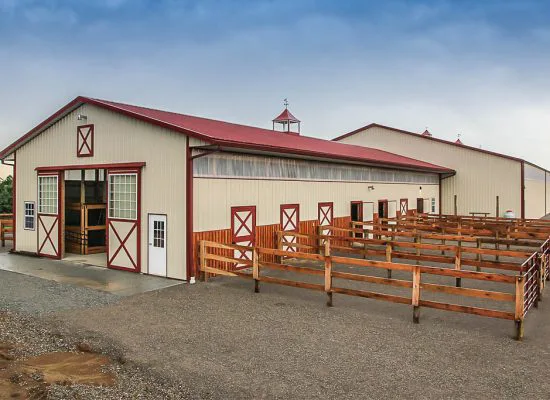 Tan horse barn with a red metal roof, red-trimmed Dutch doors, and wooden corral fencing