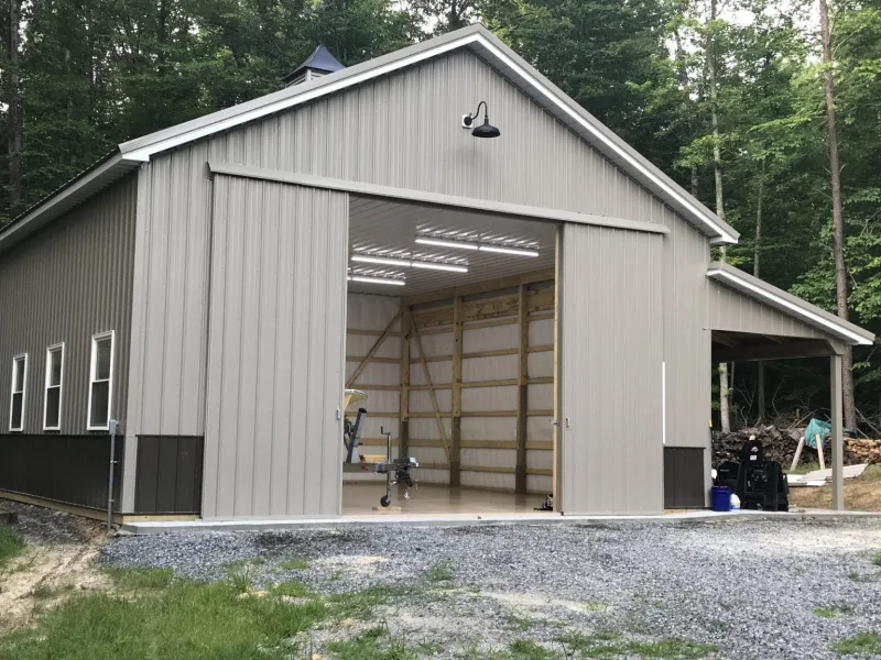Gray pole barn storage building with a wide sliding door open to a lit interior and a side lean-to