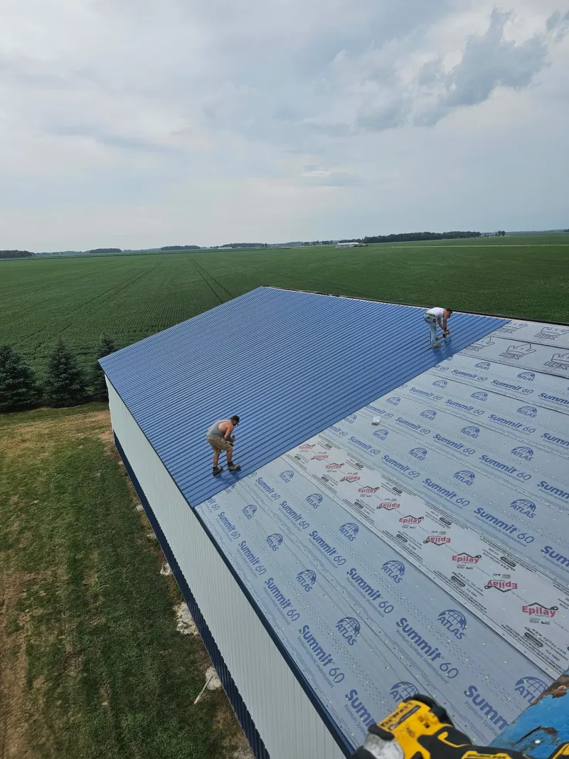 Workers installing blue metal roof panels over synthetic underlayment on a post-frame building